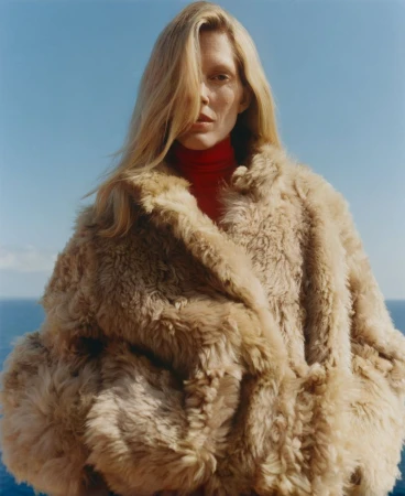Woman wearing a voluminous beige shearling coat over a red turtleneck with the blue ocean and sky in the background.