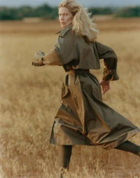 Model in an olive green belted trench coat running across a golden grassy field while looking back over her shoulder.