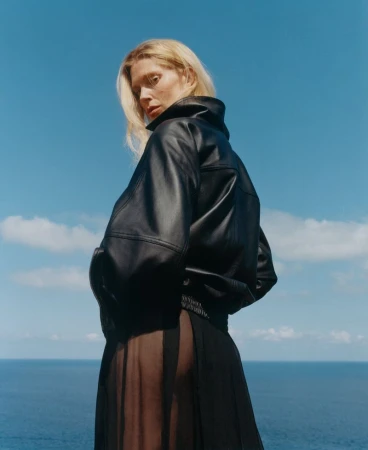 Blonde woman looking over her shoulder wearing an oversized black leather jacket and sheer skirt against an open blue ocean background.