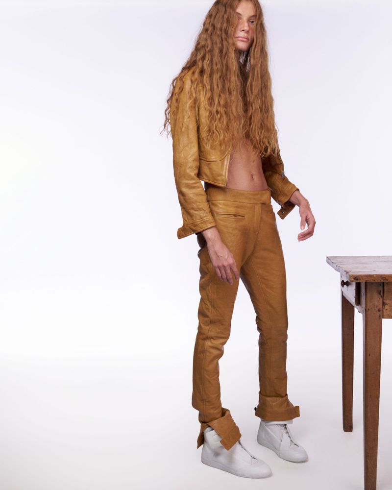 Model with long crimped hair in a tan leather suit posing beside a wooden table against a white background.