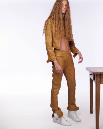 Model with long crimped hair in a tan leather suit posing beside a wooden table against a white background.