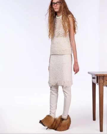 Larissa Moraes standing in a white knit top, fringed sequin skirt, and oversized fur heels against a white studio background.