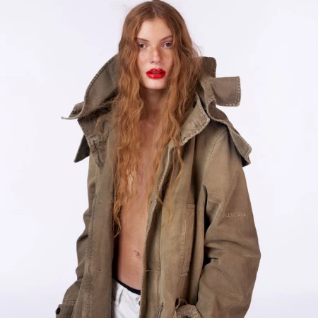Larissa Moraes wearing an oversized khaki hooded parka jacket with glossy red lipstick against a white studio backdrop.