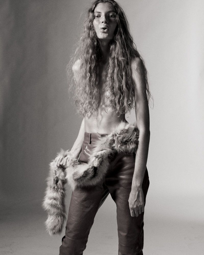 Model Larissa Moraes with long wavy hair wearing leather trousers and a fur stole against a studio backdrop.