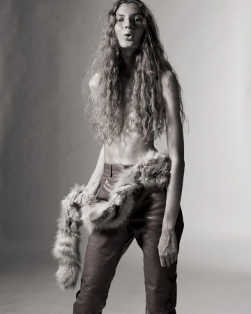 Model Larissa Moraes with long wavy hair wearing leather trousers and a fur stole against a studio backdrop.