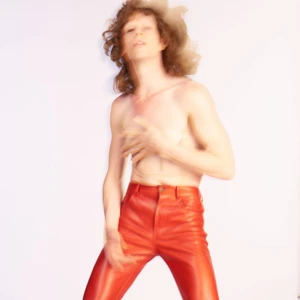 Shirtless model with shaggy brown hair wearing glossy red leather pants posed against a white studio background.