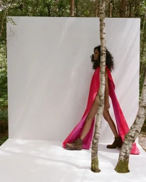 Model walking in a flowing magenta and red cape with brown combat boots across an outdoor white set in a birch forest.