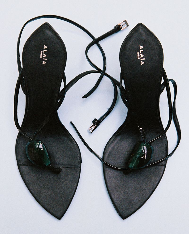Pair of black pointed-toe stiletto thong sandals with green cabochon embellishments and thin straps on a plain white surface.