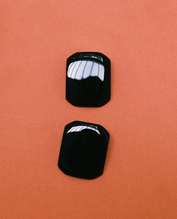 Two glossy black geometric earrings resting vertically on a smooth peach terracotta background.