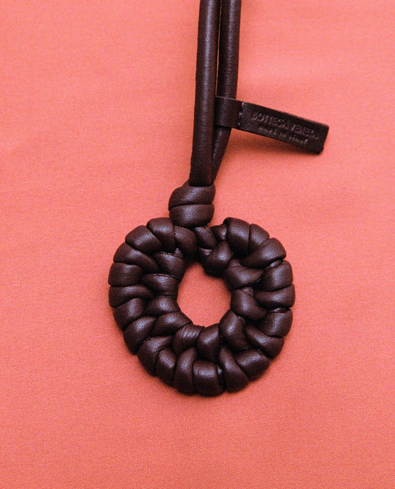 Dark brown braided leather circular pendant with a branded tag against a coral pink backdrop.