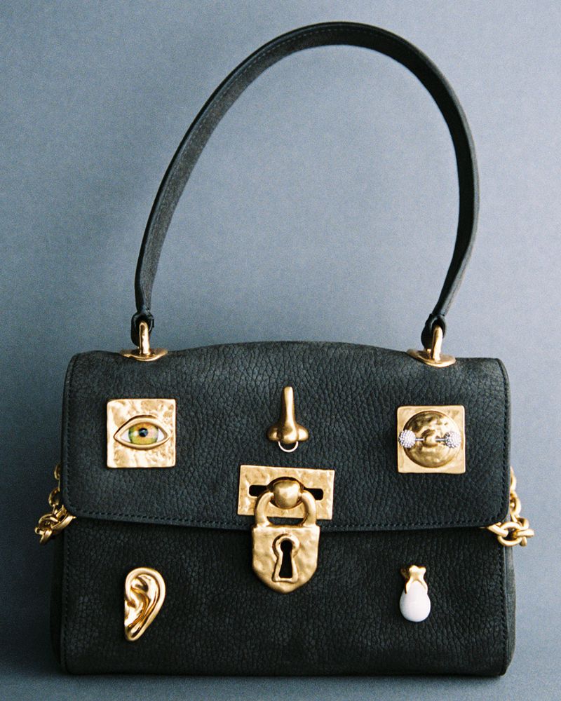 Black textured leather handbag adorned with gold surrealist face hardware including an eye, nose, and ear against a grey backdrop.
