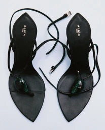 Top-down studio shot of black Alaïa pointed-toe thong heeled sandals with emerald green stones and wrap ankle ties on a white background.
