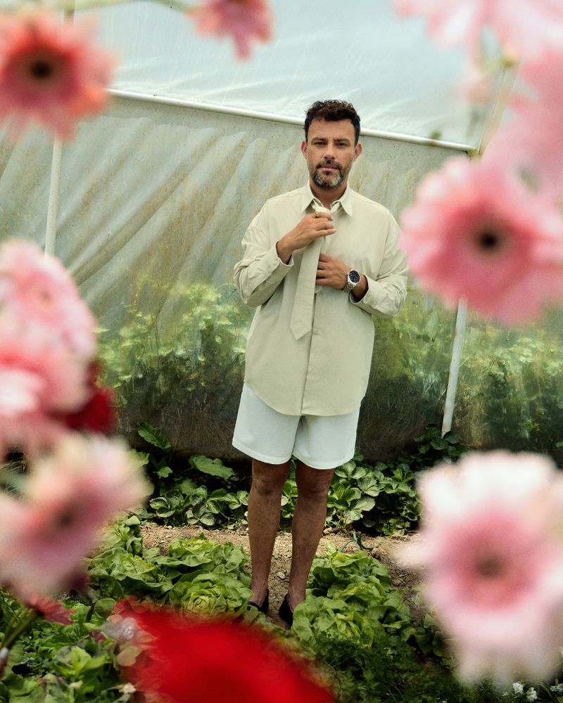 Portrait of a light-skinned adult male standing in a garden with pink flowers, wearing a beige shirt, white shorts and black dress shoes.