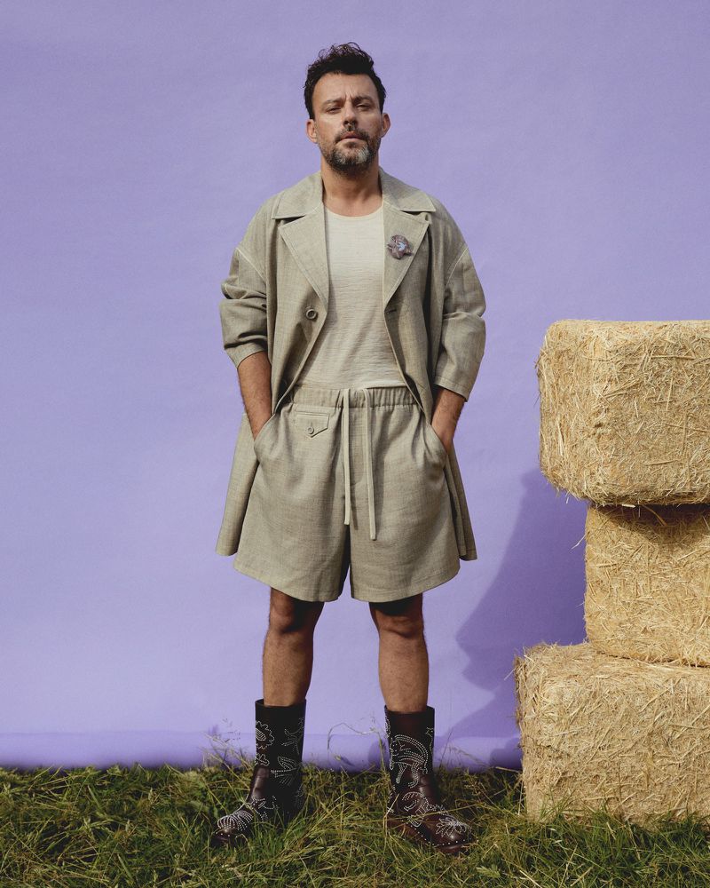 Full shot of a man in a khaki-colored short suit and cowboy boots, standing in a grassy field next to hay bales with a lavender background.