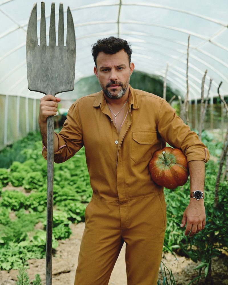 Adult male in brown jumpsuit holds large wooden garden fork and pumpkin while standing in greenhouse among rows of leafy plants.