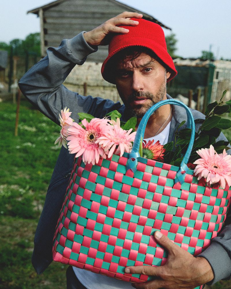 Adult male wearing red bucket hat holds a basket with pink gerbera daisies in an outdoor setting with a shed in the background.