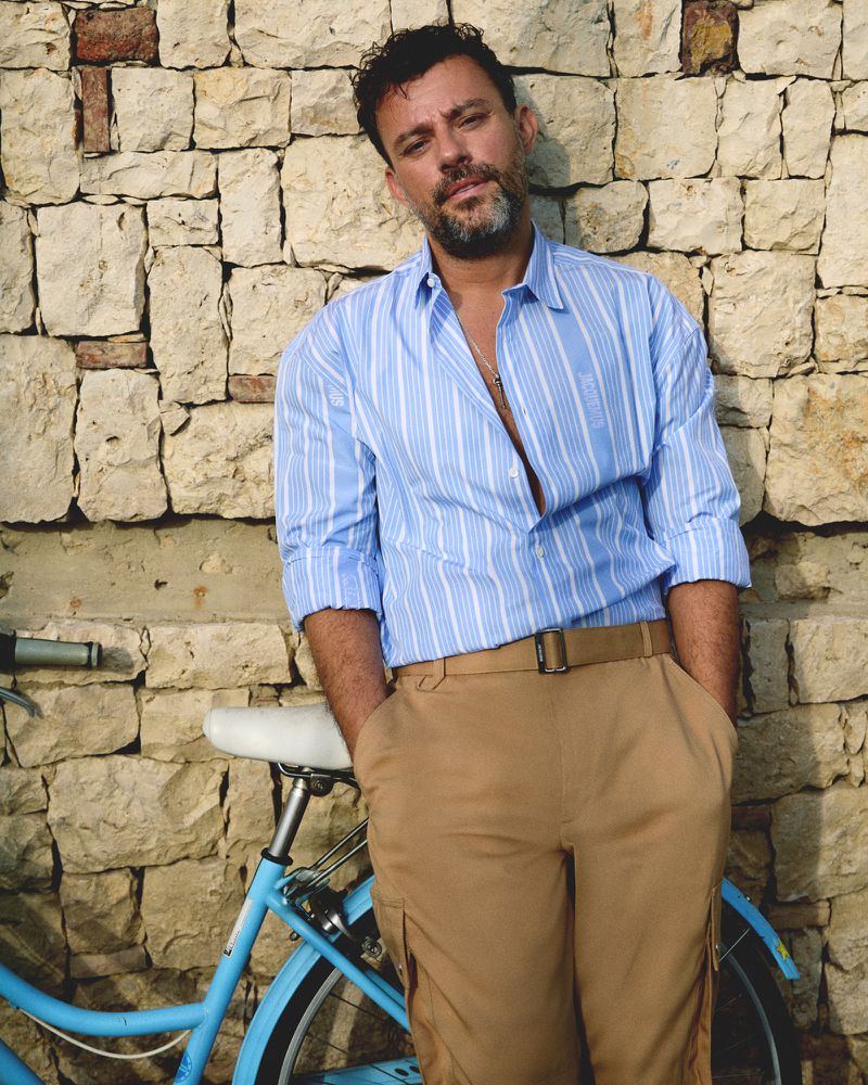 Adult man with beard in striped shirt posing against stone wall with blue bicycle in an eye-level outdoor shot. 