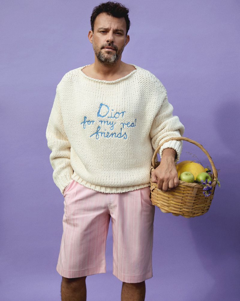 Adult male poses with a woven basket of fruit against a solid purple backdrop. He wears a large knit sweater and pink shorts.