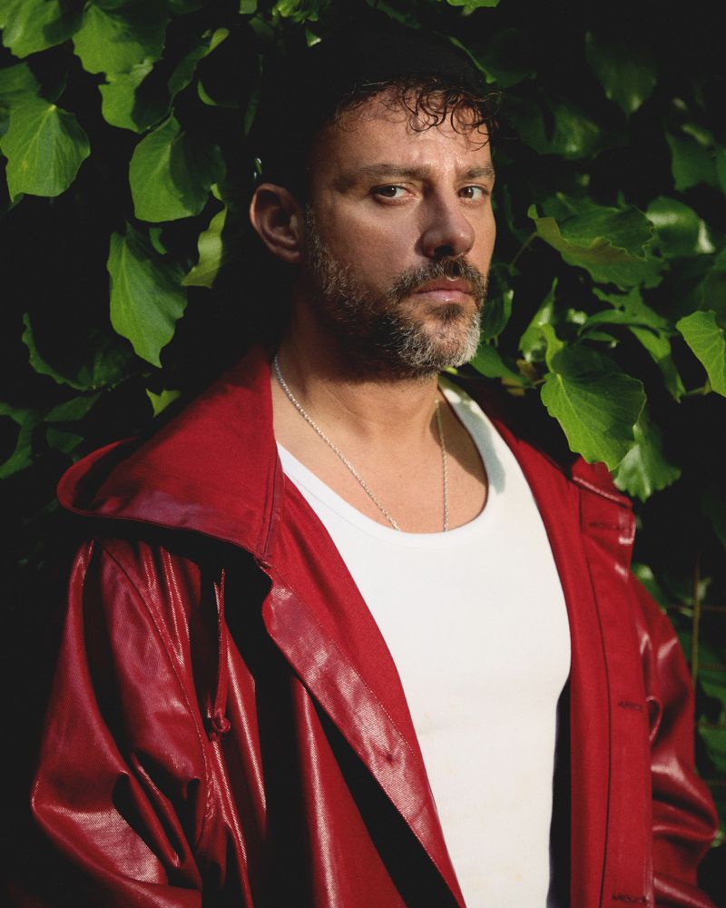 Head and shoulder shot of a man in a white t-shirt and red, shiny jacket, standing outside against a background of leafy green vines.