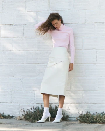 Model Rosalieke Fuchs in a pale pink turtleneck and ivory midi skirt posing against a white brick wall in white pointed heels.