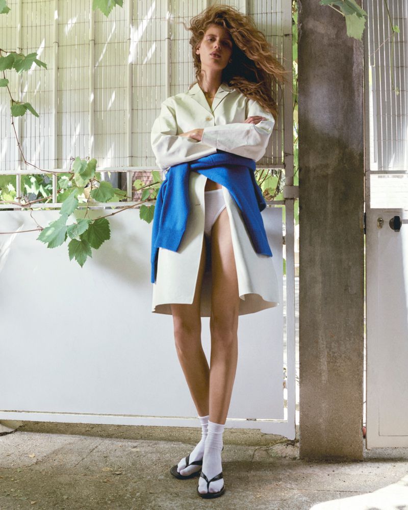 Model Rosalieke Fuchs wearing an off-white coat, blue sweater tied at the waist, white socks, and black sandals against a gate.