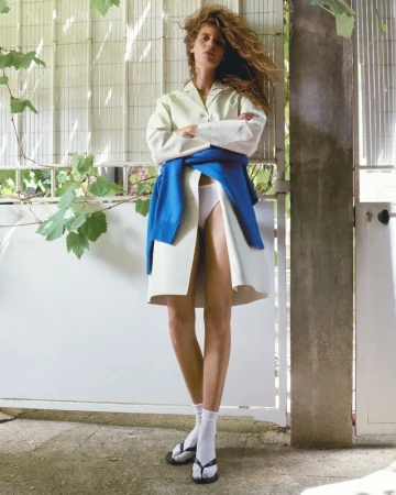 Model Rosalieke Fuchs wearing an off-white coat, blue sweater tied at the waist, white socks, and black sandals against a gate.