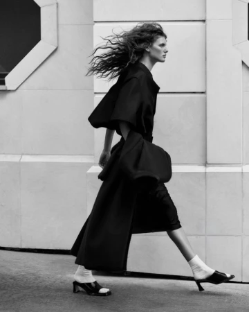 Model Rosalieke Fuchs walking outdoors in profile, wearing an oversized black coat, white socks, and black open-toe heels.