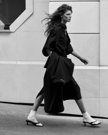 Model Rosalieke Fuchs walking outdoors in an oversized black outfit, white socks, and kitten-heel sandals by a building wall.