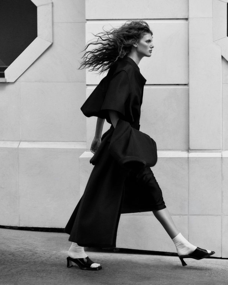 Model Rosalieke Fuchs walking past a stone architectural wall in a structured black coat, white socks, and open-toe heels.