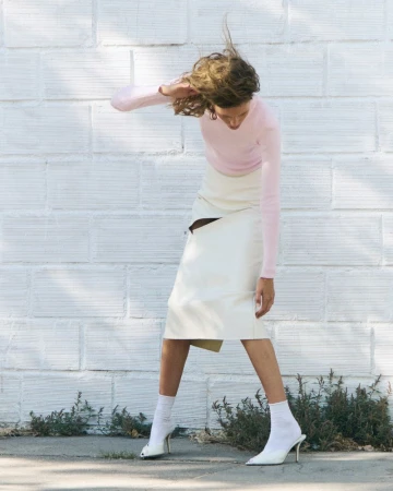 Model Rosalieke Fuchs wearing a pink long-sleeved top, cream slit midi skirt, white socks, and white heels against a white brick wall.