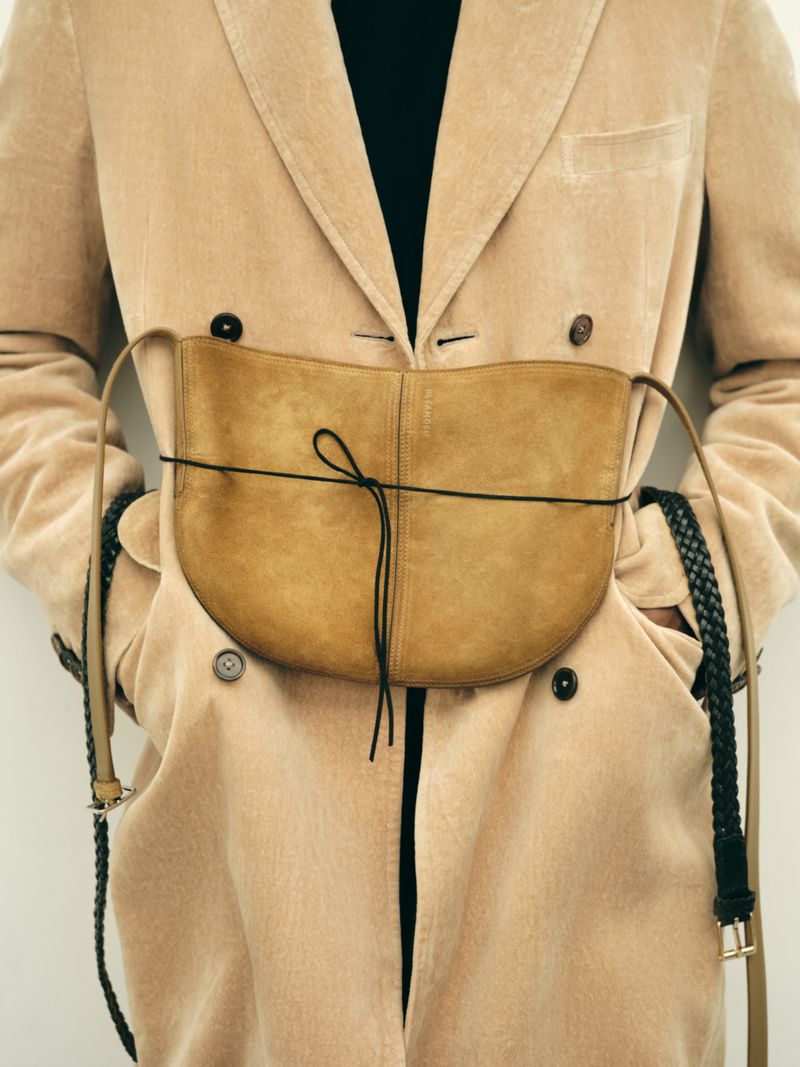 Close-up of a model wearing a beige double-breasted coat with a tan suede waist bag tied with a thin cord.