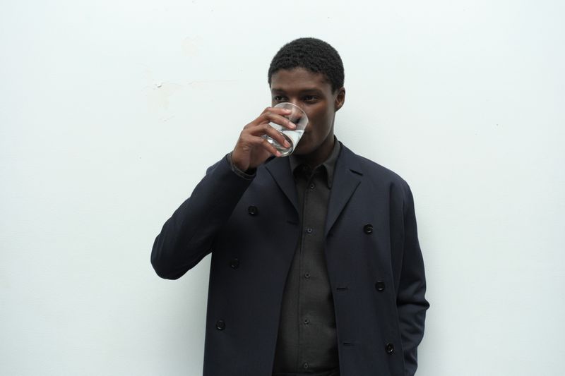 Model Aboubakar Konte wearing a dark navy coat and drinking from a glass of water against a white wall.