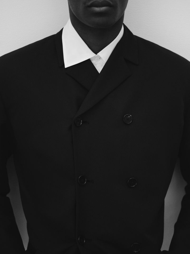 Model wearing a structured black double-breasted jacket over a crisp white collared shirt against a pale studio backdrop.