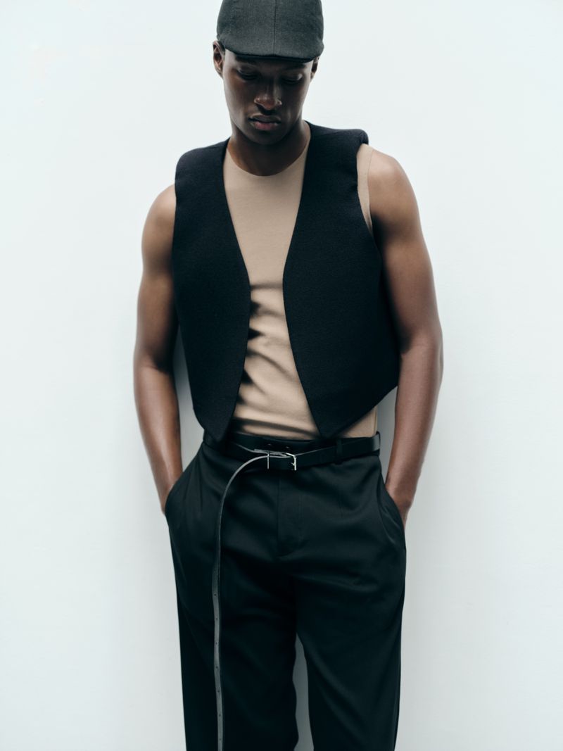 Model Aboubakar Konte wearing a black cropped waistcoat, beige sleeveless top, and flat cap against a white studio background.