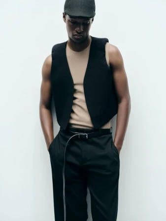 Model Aboubakar Konte wearing a black cropped waistcoat, beige sleeveless top, and flat cap against a white studio background.