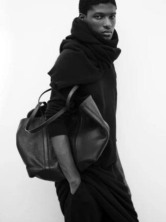 Model Aboubakar Konte posing with a large black leather tote bag and draped knit scarf against a plain white background.