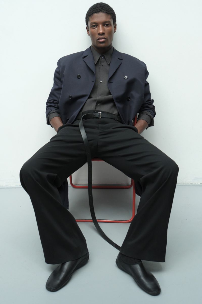 Model Aboubakar Konte seated on an orange-framed chair wearing a navy blazer, wide-leg black trousers, and a long leather belt.