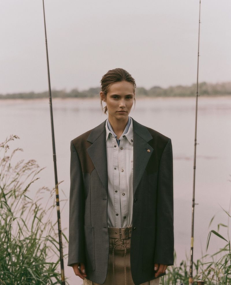 Alessandra Vo in an oversized blazer and collared shirt standing between two fishing rods by a riverbank.
