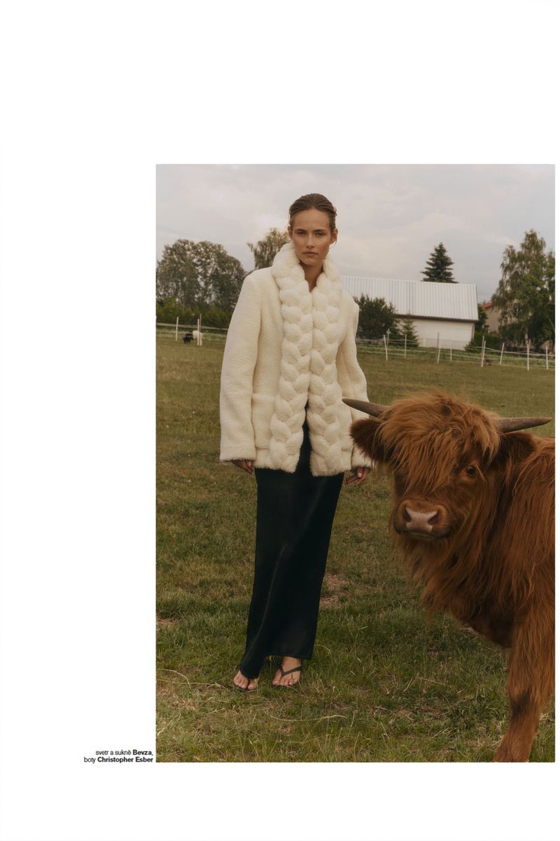 Model Alessandra Vo standing outdoors in a cream textured jacket next to a brown Highland cow in a grassy farm pasture.