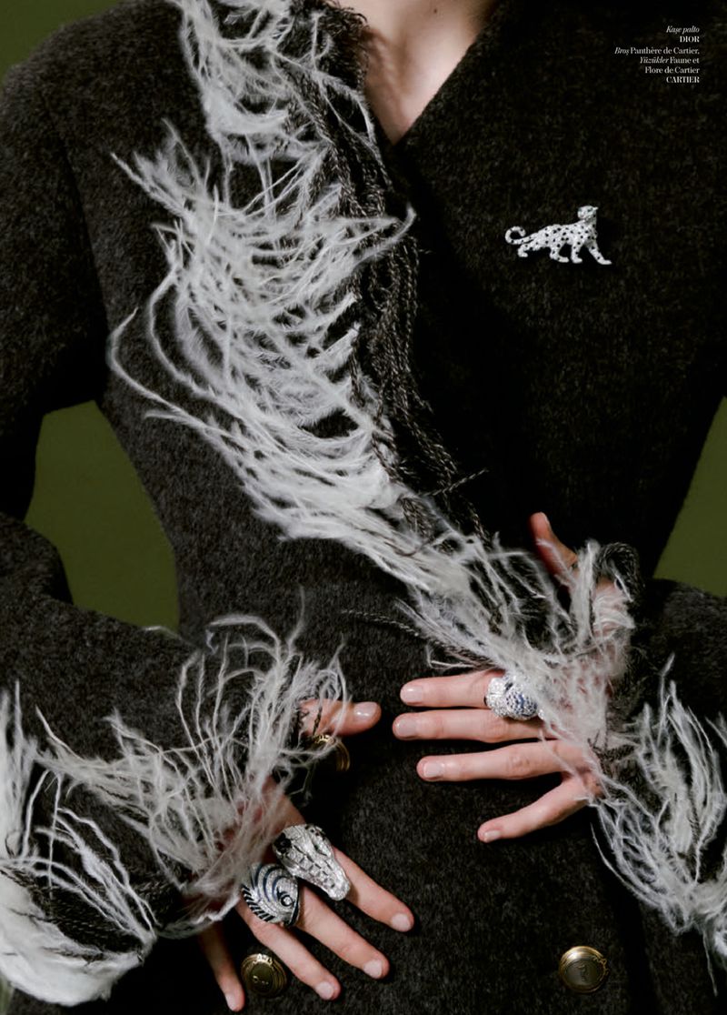 Close-up of model Patrycja Piekarska wearing a dark textured coat with white feather trim, animal brooch, and rings against green.