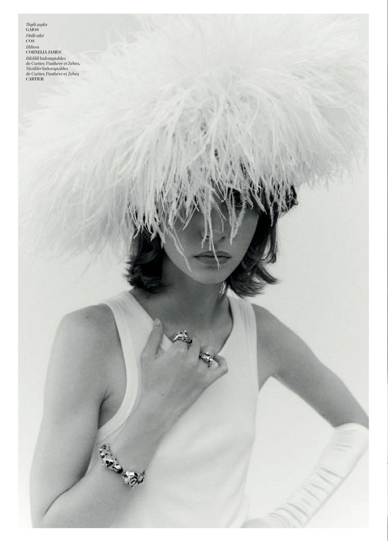 Model Patrycja Piekarska in a large white feather hat, white tank top, and opera glove against a plain white background.