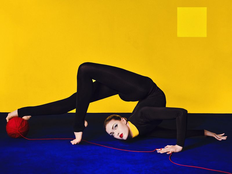 Model in a black bodysuit contorting on a blue floor with a red yarn ball against a vibrant yellow backdrop.