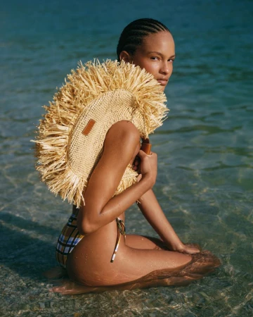 A woman in a bikini sits in shallow water holding a straw fringed bag looking back towards the camera with a serene expression.