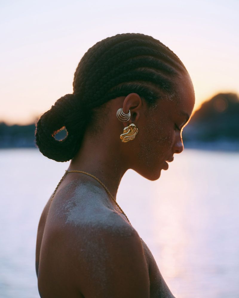 Close-up portrait of a woman with braided hair and ornate gold earrings, showcasing her natural beauty in soft, warm lighting at dusk.
