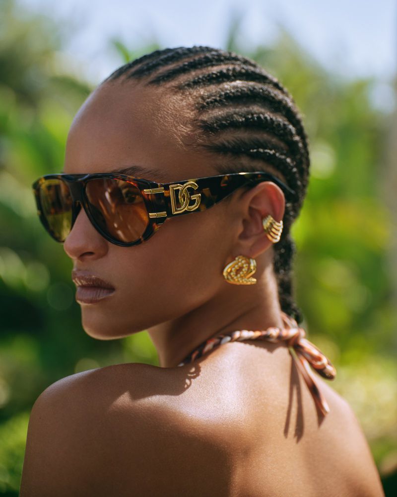 A woman with braided hair wears Dolce & Gabbana sunglasses and gold jewelry, posing outdoors with lush green foliage in the background.