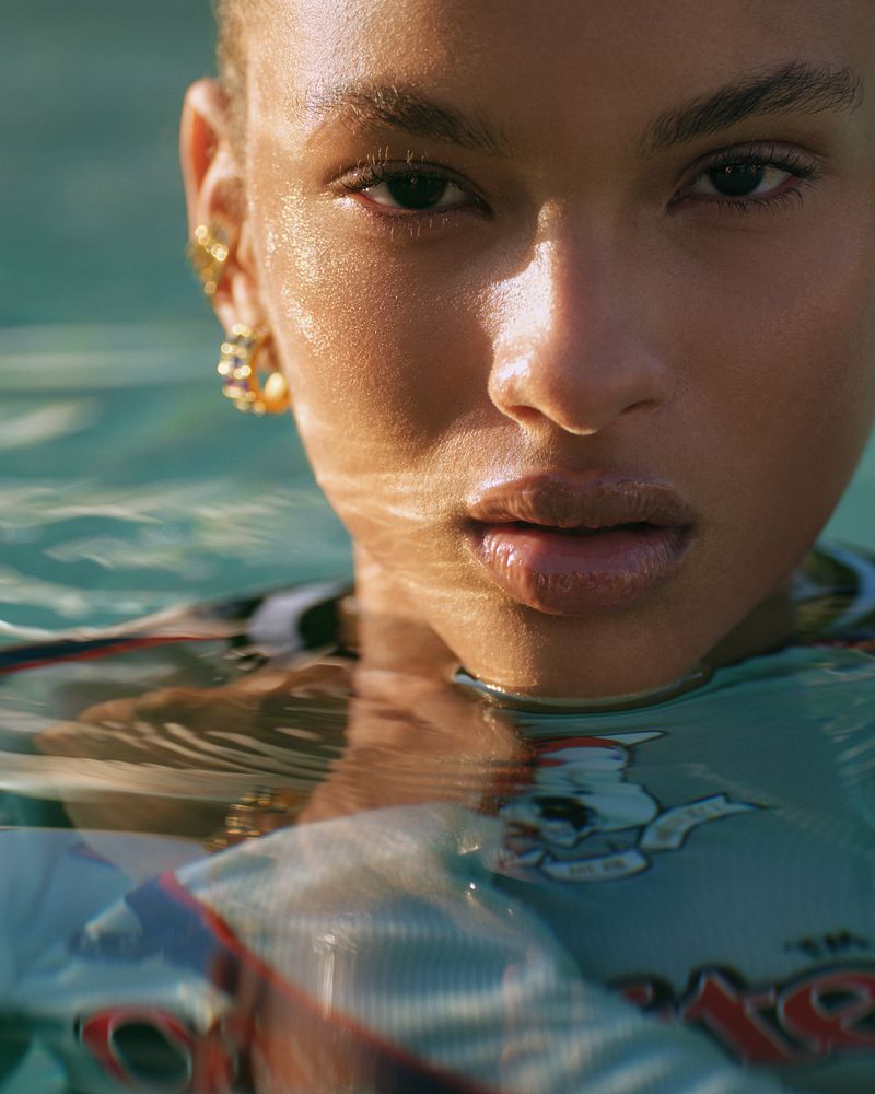 Close-up of a young adult woman in water, wearing gold hoop earrings and a printed shirt. The water is clear with slight ripples.