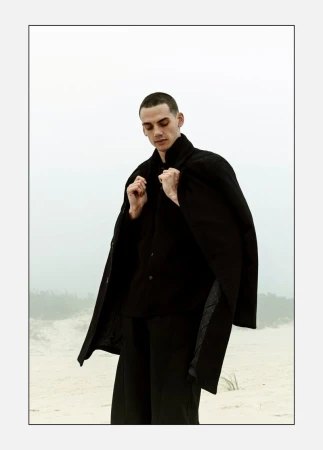 Male model in a black buttoned shirt and black overcoat holding his collar on a misty sand dune.