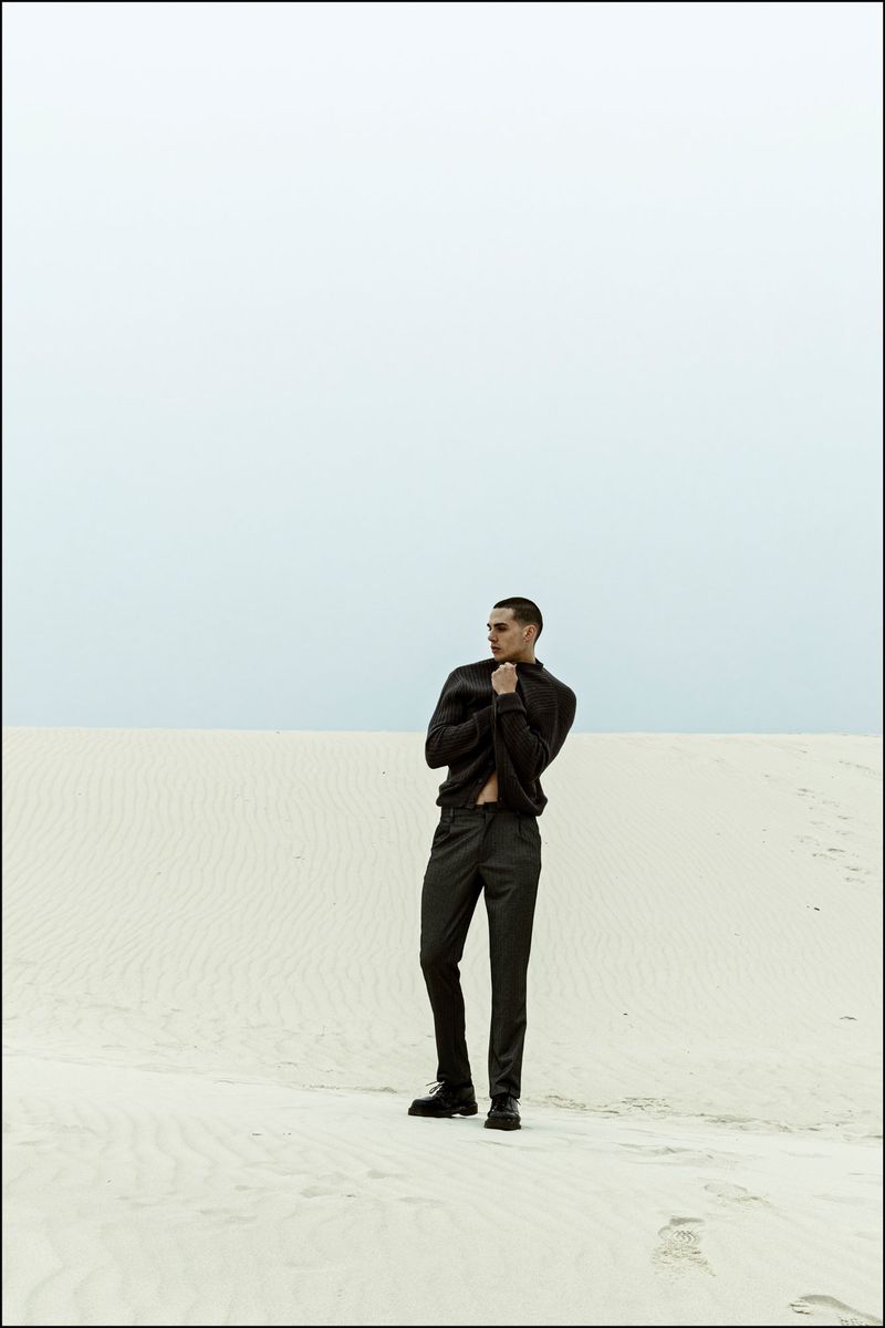 A man in a dark jacket and trousers stands on white sand dunes beneath an overcast sky.