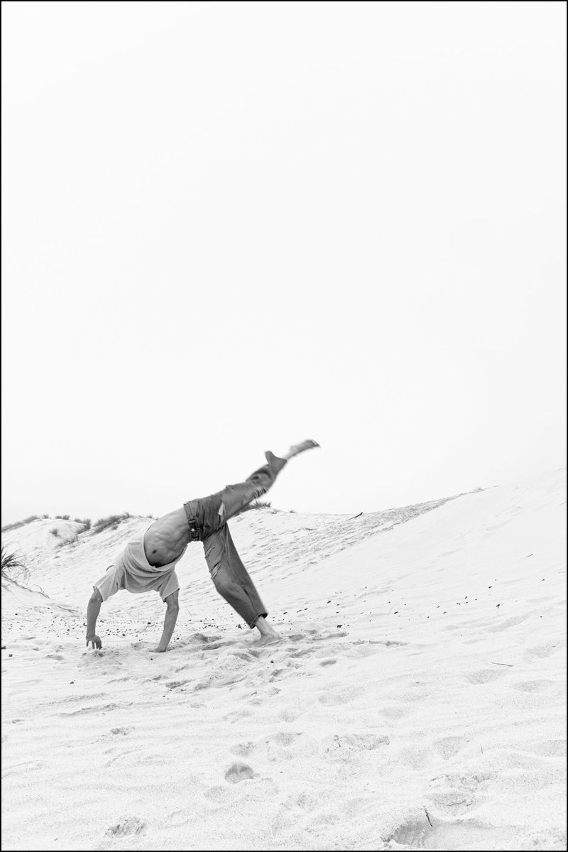 Black and white photo of a man performing an acrobatic backbend on a sand dune beneath an expansive bright sky.