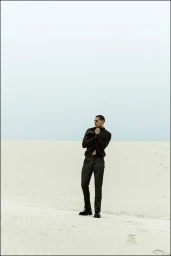 Male model in an all-black outfit standing on a white sand dune under a misty overcast sky.